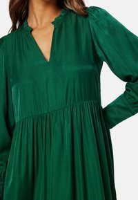 Grön, långärmad klänning med V-ringning och volangkrage, med en samlad midja och slät, glänsande tygtextur.
