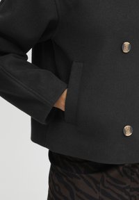Černá zkrácená bunda z hladké tkaniny, s dvouřadým zapínáním, zlatými knoflíky a bočními kapsami.