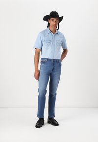 Camicia di jeans azzurro chiaro con due tasche sul petto, cappello da cowboy nero, jeans blu e scarpe nere, su sfondo bianco.