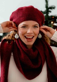 Boina y bufanda de punto en color burdeos con una textura; usando pendientes en forma de orbe plateados; sonriendo frente a un fondo navideño difuminado.