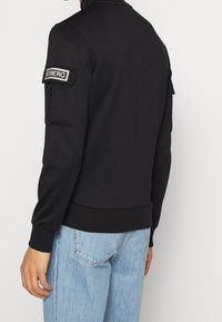 Sudadera negra con cuello, puños ajustados y dobladillo acanalado. Presenta un parche en la manga izquierda. Combinada con jeans de color azul claro.