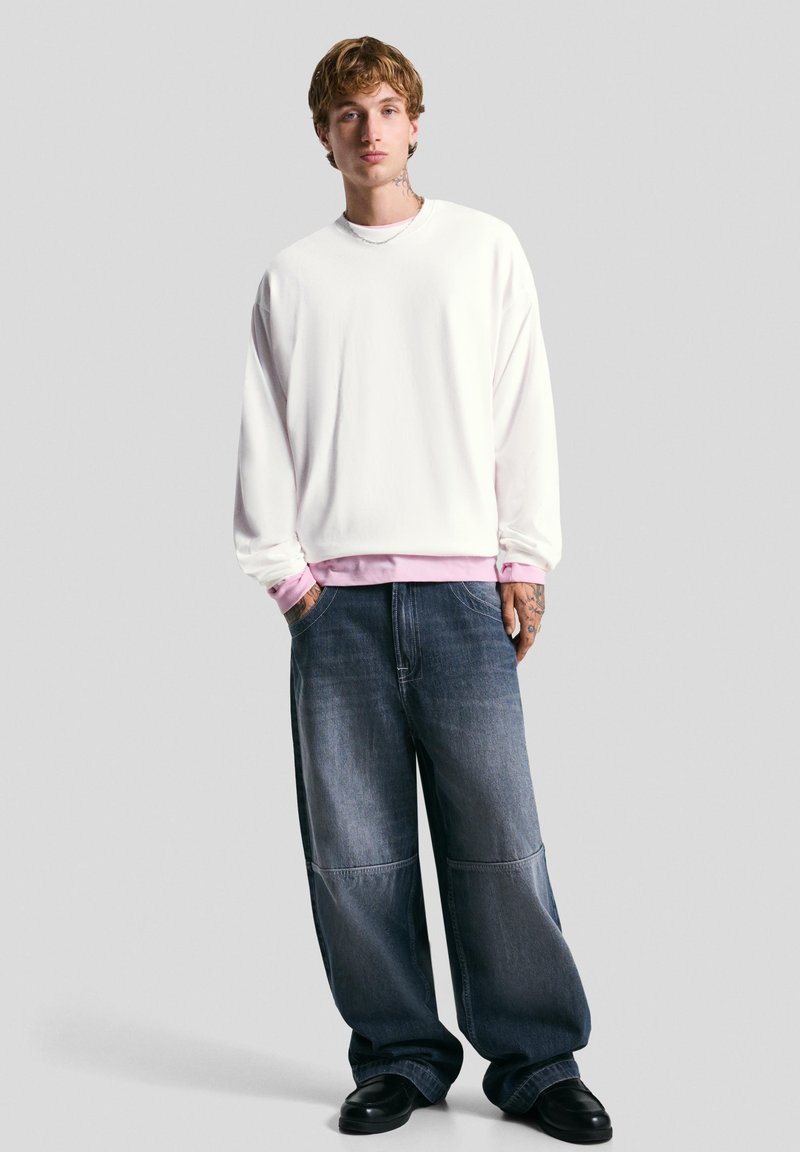 Jeune homme avec des tatouages portant un sweat-shirt blanc sur une chemise rose, un jean bleu ample et des chaussures noires, se tenant devant un fond uni.