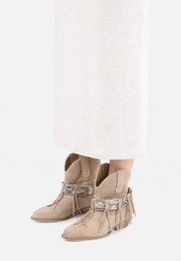 Suede ankle boots in beige, featuring fringe accents and metallic detail at the ankle. Pointed toe and low stacked heel design.