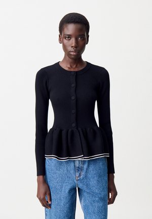 Femme portant un cardigan noir ajusté à côtes avec des boutons et un ourlet en peplum, associé à un jean en denim bleu taille haute, se tenant devant un fond blanc.