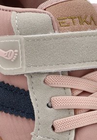 Zapatilla de color rosa claro con detalles texturizados en gris, que presenta una correa de velcro, cordones rosas y una franja lateral de color azul oscuro. Logo en dorado.