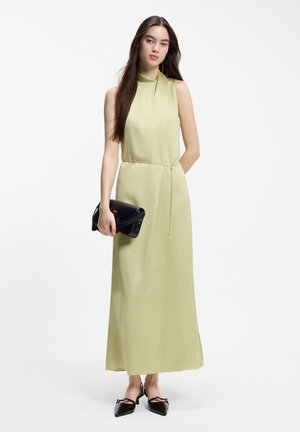 Femme aux longs cheveux foncés portant une robe longue vert clair sans manches, des ballerines pointues noires, et tenant une pochette noire sur fond blanc.