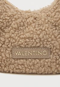 Beige teksturēta soma ar pūkainu, maigu virsmu un izteiksmīgu zeltainu "VALENTINO" logotipu uz ādas plāksnītes.