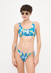 Kék virágmintás bikini, fehér és sárga mintákkal. Cropped fazonú felsője széles pántokkal és oldalt megkötős alsója van. Texturált anyag.