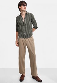 Camicia a righe verde oliva con maniche arrotolate, abbinata a pantaloni béige con pieghe e mocassini marroni. Inclusi occhiali da sole trasparenti.