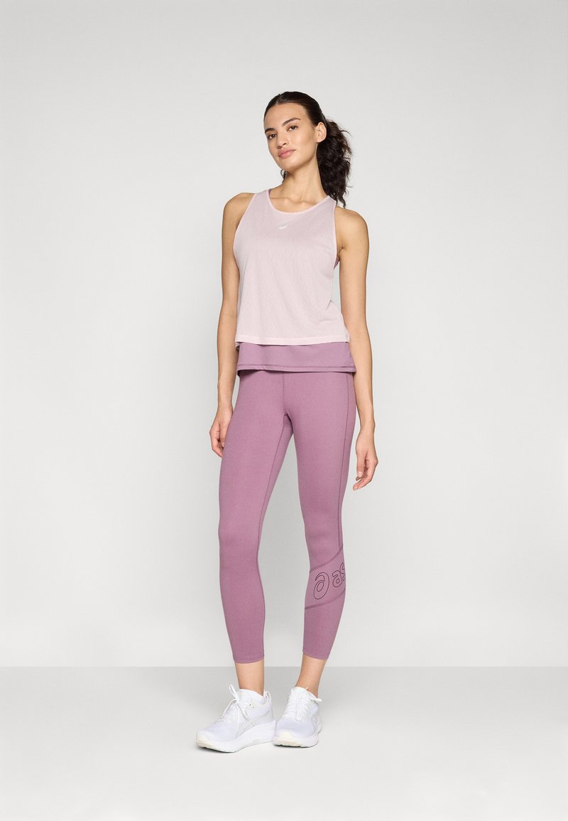Un débardeur rose clair superposé sur un legging lilas ajusté avec un logo en relief. Le modèle porte des chaussures de sport blanches sur un fond neutre.