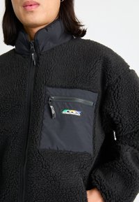 Schwarze Fleecejacke mit hohem Kragen, ausgestattet mit einer markanten, mit Reißverschluss versehenen Brusttasche und einem mehrfarbigen Logo. Weicher, strukturierten Stoff.