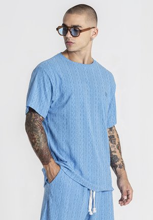 Homme tatoué portant une chemise bleu clair texturée oversize et un short assorti, des lunettes de soleil marron et des boucles d'oreilles, posant avec une main dans la poche.
