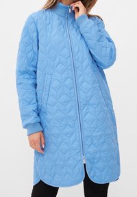 Ljusblå kviltad jacka med hög krage, dragkedja fram och ribbade ärmslut. Har sidofickor och rundad nederkant med tryckknappar.