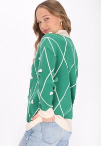 Maglione verde con un motivo a rombi, caratterizzato da dettagli bianchi e pom-pom. Polsini e orlo a coste in crema, con una vestibilità comoda.