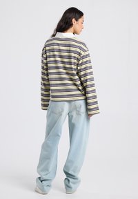 Nainen, jolla on tummat hiukset, pukeutuneena kelta-ruskearaidalliseen pitkähihaiseen paitaan, vaaleisiin löysiin farkkuihin ja valkoisiin kenkiin, seisoo selkä kameraan päin.