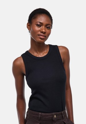 Femme aux cheveux courts porte un haut noir sans manches et un pantalon foncé, debout devant un fond clair et uni, regardant l'appareil photo.