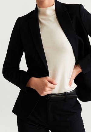 Person wearing a black blazer over a white turtleneck sweater, with black pants and a black belt, hands adjusting the blazer and waistband.