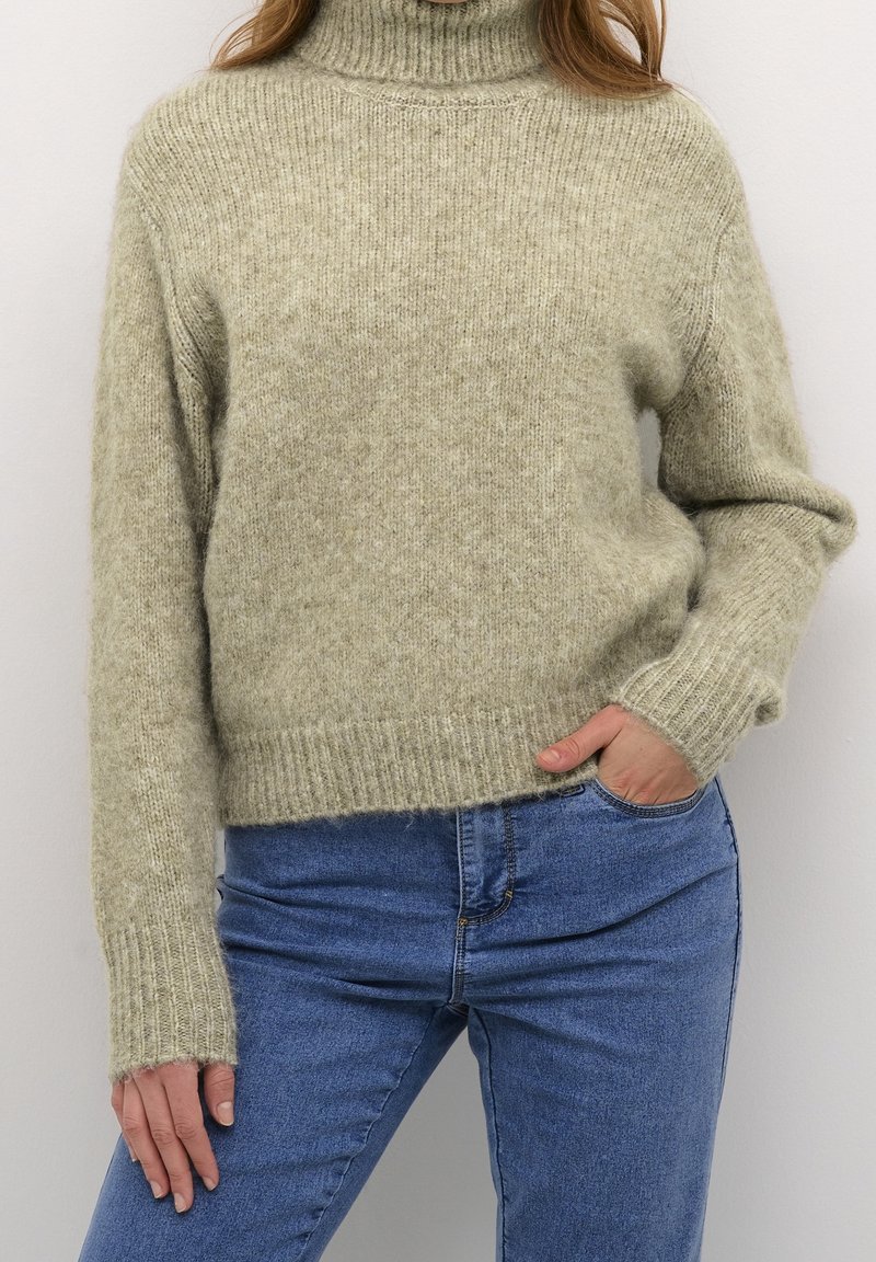 Femme portant un pull à col roulé en maille gris clair et un jean bleu, une main dans la poche, sur fond blanc.