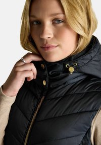 Jauna sieviete ar blondiem matiem piestiprina melnā quiltētā vesta apkakli ar zelta rāvējslēdzēju un pogām, uzvilkusi bēša garo piedurkņu krekli.