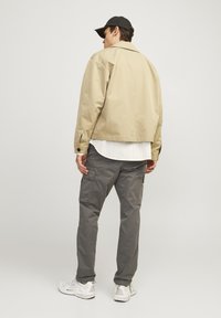 Beige bomberjacka med en cropped design, featuring en spridd krage och bruna knappdetaljer, bärs över en vit texturerad skjorta och grå cargobyxor.
