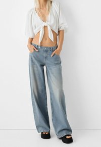 Kvinna i en vit kort topp med knytning framtill, ljusblå vida jeans, svarta plattformssandaler, poserar med händerna i jeansfickorna.