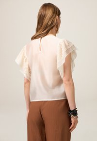 Chemisier blanc avec des détails en dentelle, manches courtes à volants ; ouverture en forme de clé au dos ; associé à un pantalon large en marron texturé.