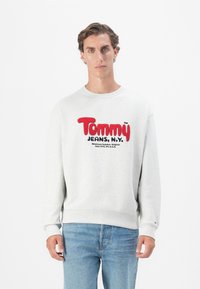 Gray pullover sweatshirt with a round neckline, featuring bold red and black "Tommy Jeans, N.Y." logo. Ribbed cuffs and hem. Blue jeans.