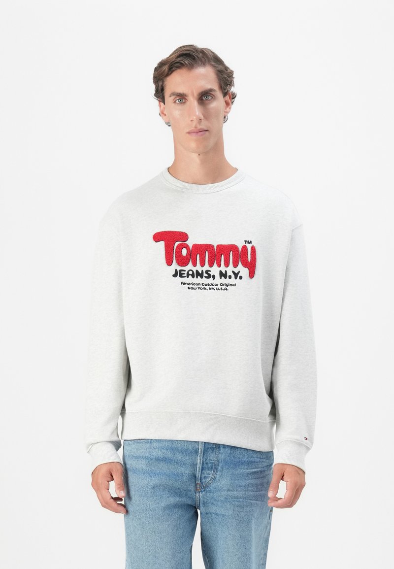 Gray pullover sweatshirt with a round neckline, featuring bold red and black "Tommy Jeans, N.Y." logo. Ribbed cuffs and hem. Blue jeans.
