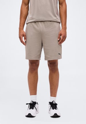 Mężczyzna w taupe krótkich spodenkach z logo Puma, białych skarpetkach i białych trampkach z czarnymi sznurówkami stoi na białym tle.