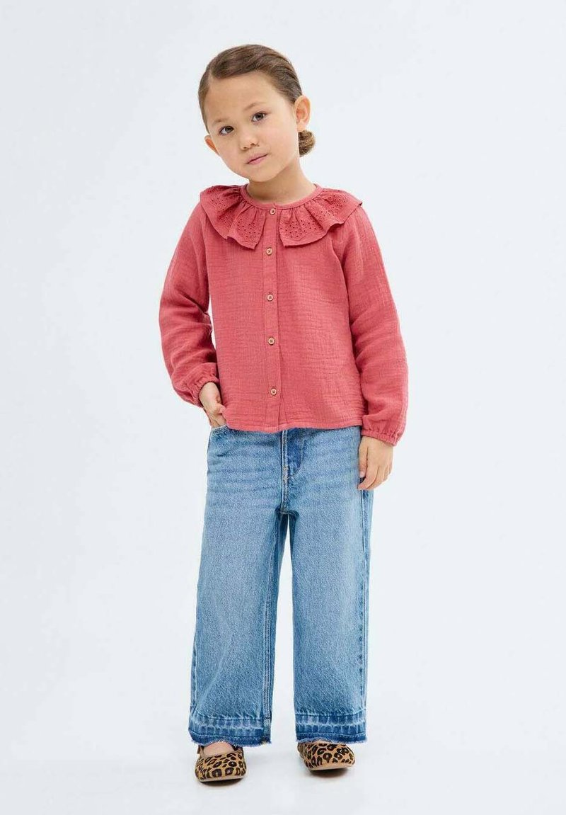 Lapsi seisoo itsevarmasti vaaleanpunaisessa paidassa, jossa on röyhelökaulus, yhdistettynä löysiin farkkuihin ja eläinkuosilla varustettuihin kenkiin, neutraalin taustan edessä.