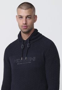 Marineblaue Kapuzenjacke mit gestricktem Muster, ausgestattet mit einem hohen Kragen, Zugbändern und dem gestickten Logo "KORUSHI ESTABLISHED 2001" auf der Vorderseite.