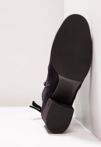 Bottine en daim noir avec détail zippé, semelle et talon vers l'avant, sur fond de surface blanche en angle.