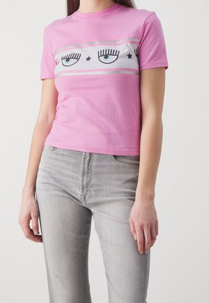 Kobieta ubrana w różowy T-shirt z krótkim rękawem z nadrukowanymi rzęsami i oczami na klatce piersiowej, w połączeniu z jasnoszarymi jeansami.