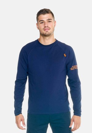Jonge man die een marineblauw shirt met lange mouwen draagt met oranje tekst op de mouw en een donkere broek, staand tegen een effen lichte achtergrond.