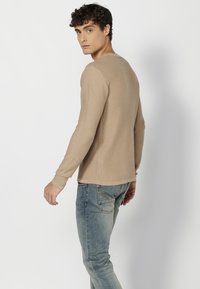 Beige színű, hosszú ujjú ing, hullámos textúrával, kerek nyakkivágással és testhez álló ujjakkal, világoskék farmernadrággal és slim fit szabással párosítva.