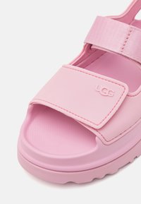 Roze gumene sandale s širokim, podesivim remenom i teksturiranim uloškom; UGG logo utisnut na remenu. Debeli, rebrasti potplat.