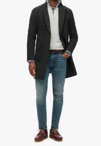 Manteau gris foncé croisé porté sur un pull gris clair, jean bleu et chaussures bordeaux. Comprend une chemise à carreaux en dessous.