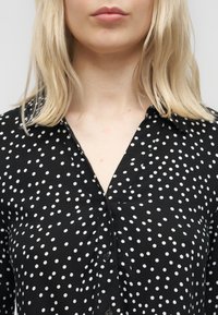 Ξανθό άτομο που φοράει μαύρο πουκάμισο με γιακά και διασκορπιστές λευκές πουά, εμφανιζόμενο από τους ώμους έως τα χείλη σε απλό φόντο.