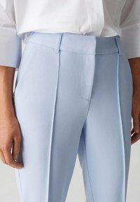 Helle blaue maßgeschneiderte Hosen aus glattem Stoff, mit flachem Frontbereich, seitlichen Taschen und dezenten Stickdetails.