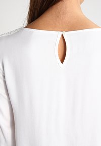 Blusa bianca con un'apertura a chiave sul retro, dotata di una piccola chiusura con bottone. Tessuto liscio con dettagli di texture minimi.