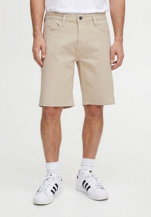 Mężczyzna w beżowych, sięgających do kolan szortach, białej koszulce, białych skarpetkach i białych butach Adidas z czarnymi paskami, stojący na tle jednolitego tła.