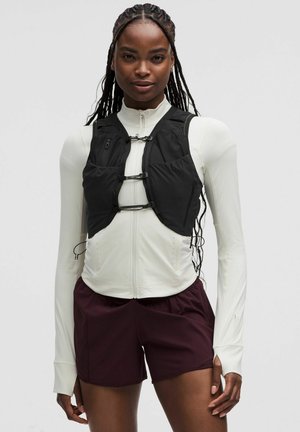 Sort multifunktionel vest med justerbare stropper, lynlåslommer og letvægtsmateriale, båret over en hvid langærmet top og maroon shorts.