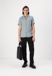 Camisa de botones de manga corta, de color azul claro, con un bolsillo en el pecho, combinada con pantalones negros y una bolsa de tote negra, llevada con zapatos negros.
