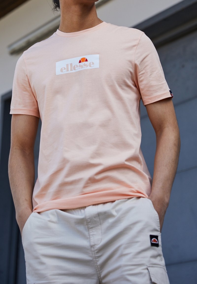 Světle broskvové bavlněné tričko s bílou nášivkou loga a červenými a oranžovými akcenty. Nosit s béžovými šortkami, ruce v kapsách.