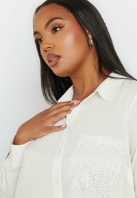 Camicia bianca con bottoni, dotata di una tasca testurizzata adornata da decorazioni in argento. Tessuto liscio e design del collo classico.