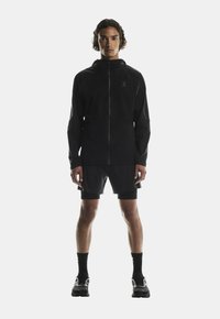 Giacca impermeabile nera con chiusura a zip, maniche corte e cappuccio. Abbinata a pantaloni corti neri e scarpe, presenta un design atletico elegante.
