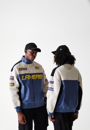 Chaqueta de los Lakers con un diseño en crema, azul y negro, con grandes logotipos, cierre con cremallera en la parte delantera y puños acanalados. Llevada por dos personas, vista desde el frente y desde la parte trasera.