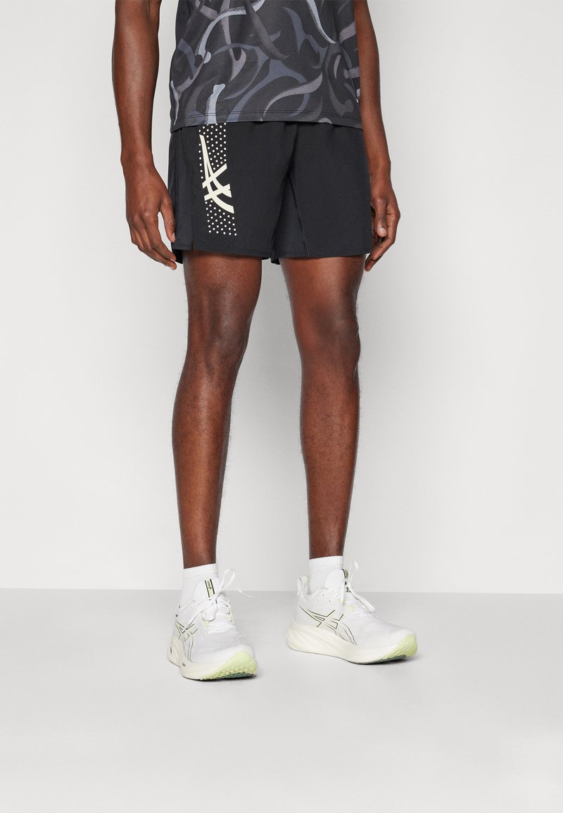 Pantalones cortos deportivos negros con un acento estampado y puntos blancos en un lado, combinados con zapatillas blancas con suelas verdosas.