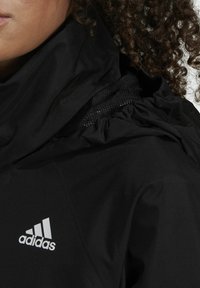 Giacca nera con cappuccio e tessuto arricciato intorno al collo, con mesh nera sotto e un logo Adidas bianco nella zona del petto.