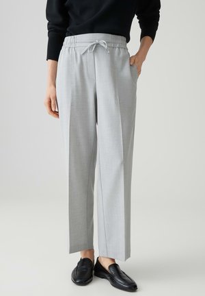 Personne portant un pantalon gris clair à jambes droites avec taille à cordon et des mocassins noirs, debout, une main dans la poche.